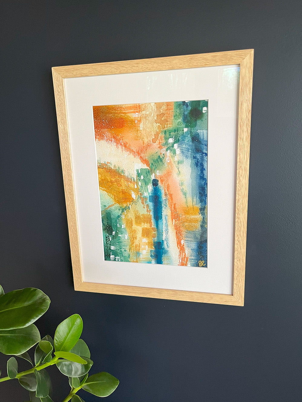 Framed Watercolour Abstract called Yearning hanged on a dark blue wall with plant